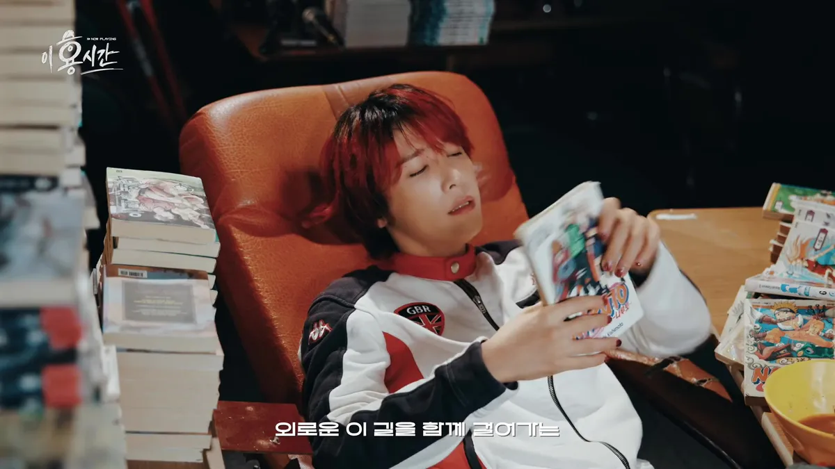 Lee Hong-gi canta mientras sostiene un manga sentado en una silla