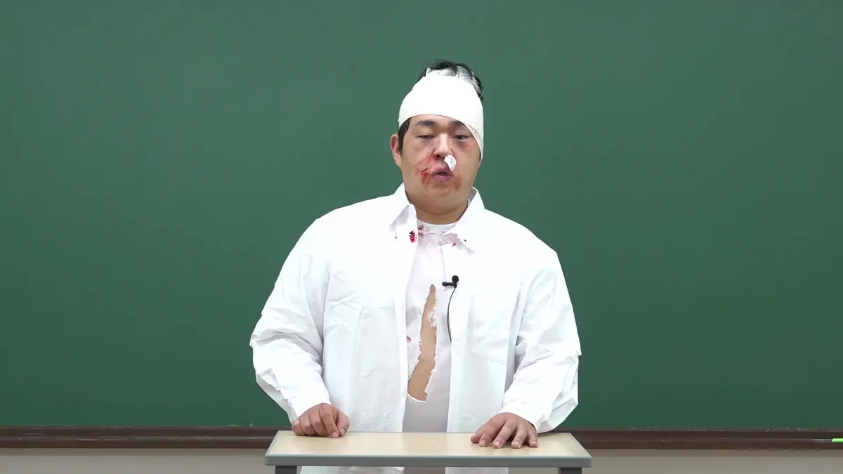頭に包帯を巻き、血の付いた服を着たスミンが黒板の前に座っている