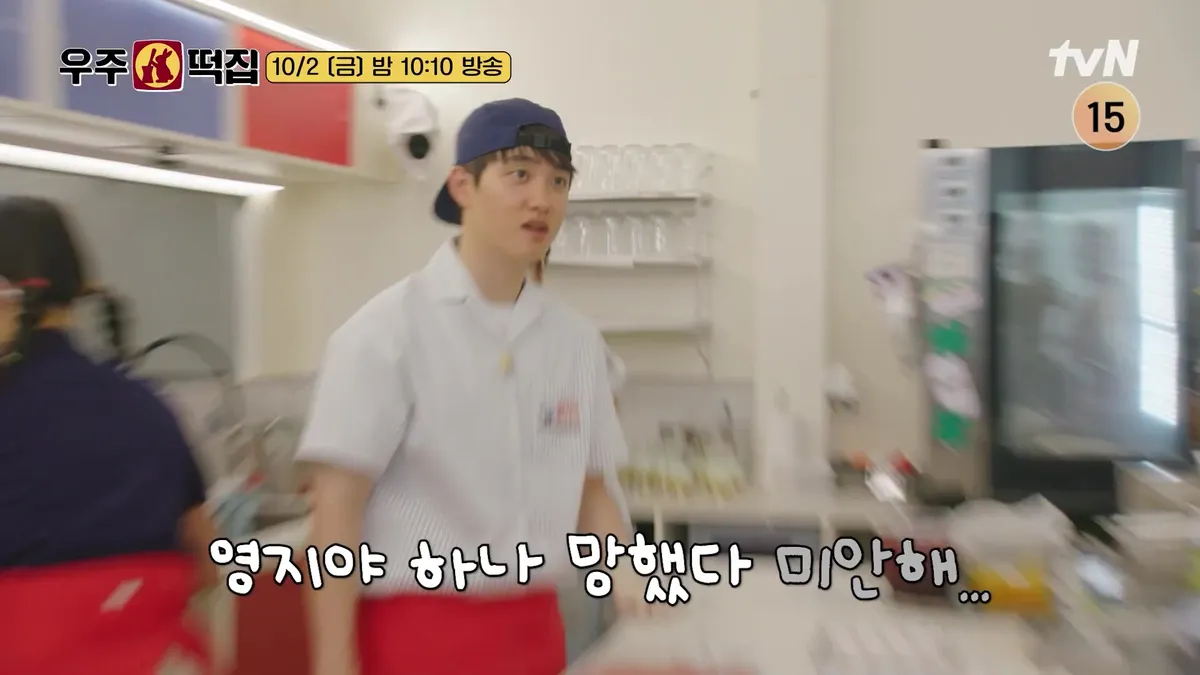 D.O. stands wearing a white shirt and a blue hat